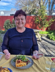 A smiling Chandra sitting at an outdoor table, holding a small glass bowl in her left hand, before it was broken. She is wearing a navy-blue top and a glass pendant necklace. In front of her is a plate with a slice of quiche and a fresh green salad.  