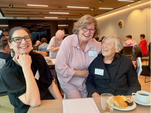 Three women sitting at a table with coffee and cake all smiling for the camera at the reception after the Access Matters summit.