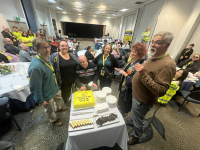 Kevin about to cut the cake. The cake is square and has bright yellow icing. Several other people are standing around the table