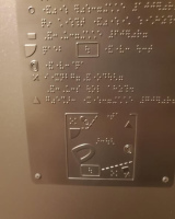 Close-up of a metal panel with raised braille text and tactile symbols, including simple shapes and a small layout map, likely used as an accessible guide on a train or in a public space.