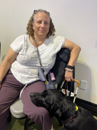 Sally is sitting on a chair with her new guide dog Priya leaning on her knee looking at her