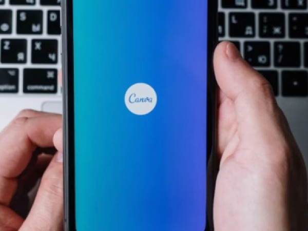 Close-up of a smartphone held in both hands, displaying the Canva app logo on a blue gradient screen, with a laptop keyboard blurred in the background. 