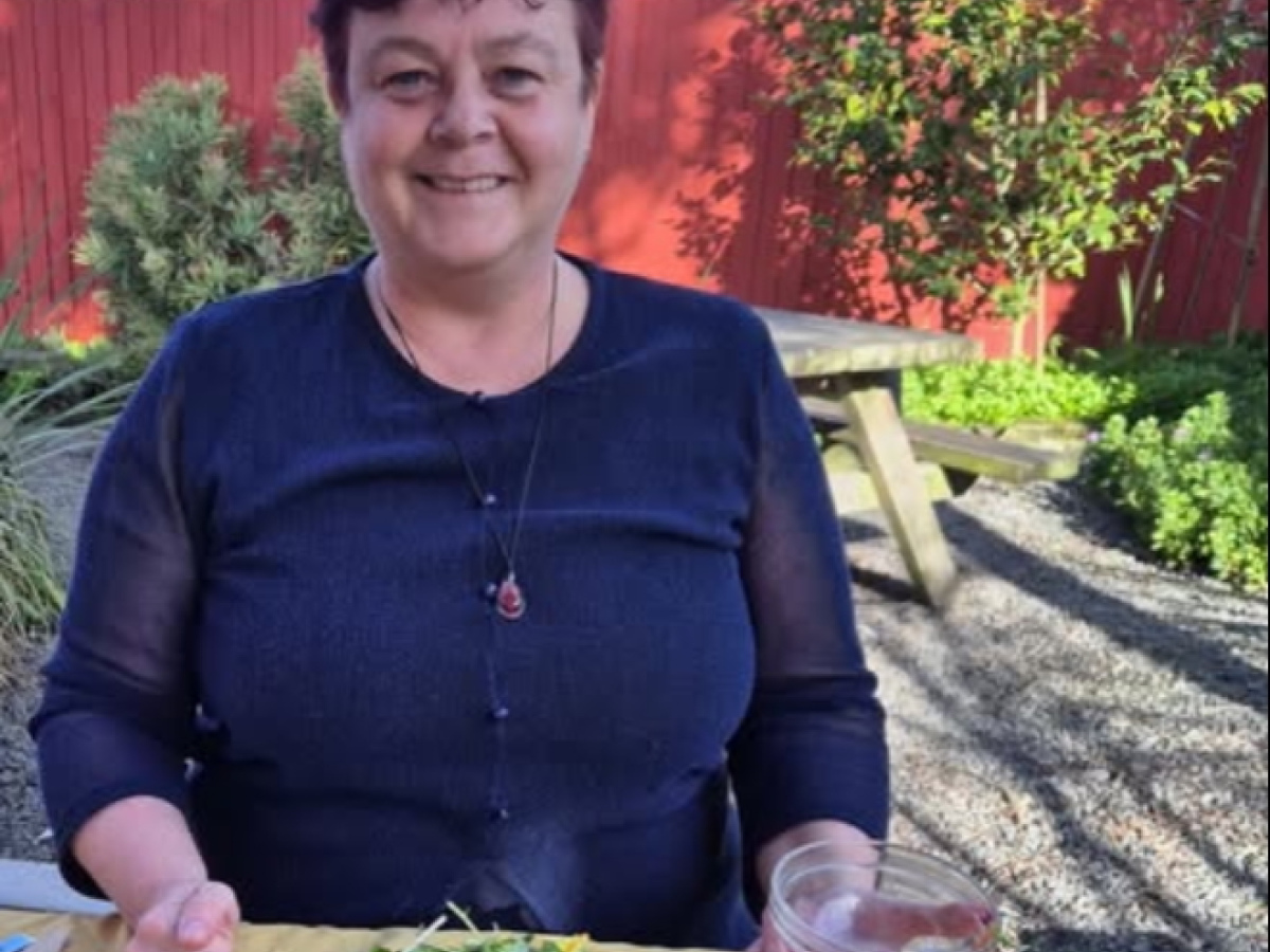 A smiling Chandra sitting at an outdoor table, holding a small glass bowl in her left hand, before it was broken. She is wearing a navy-blue top and a glass pendant necklace. In front of her is a plate with a slice of quiche and a fresh green salad. The t
