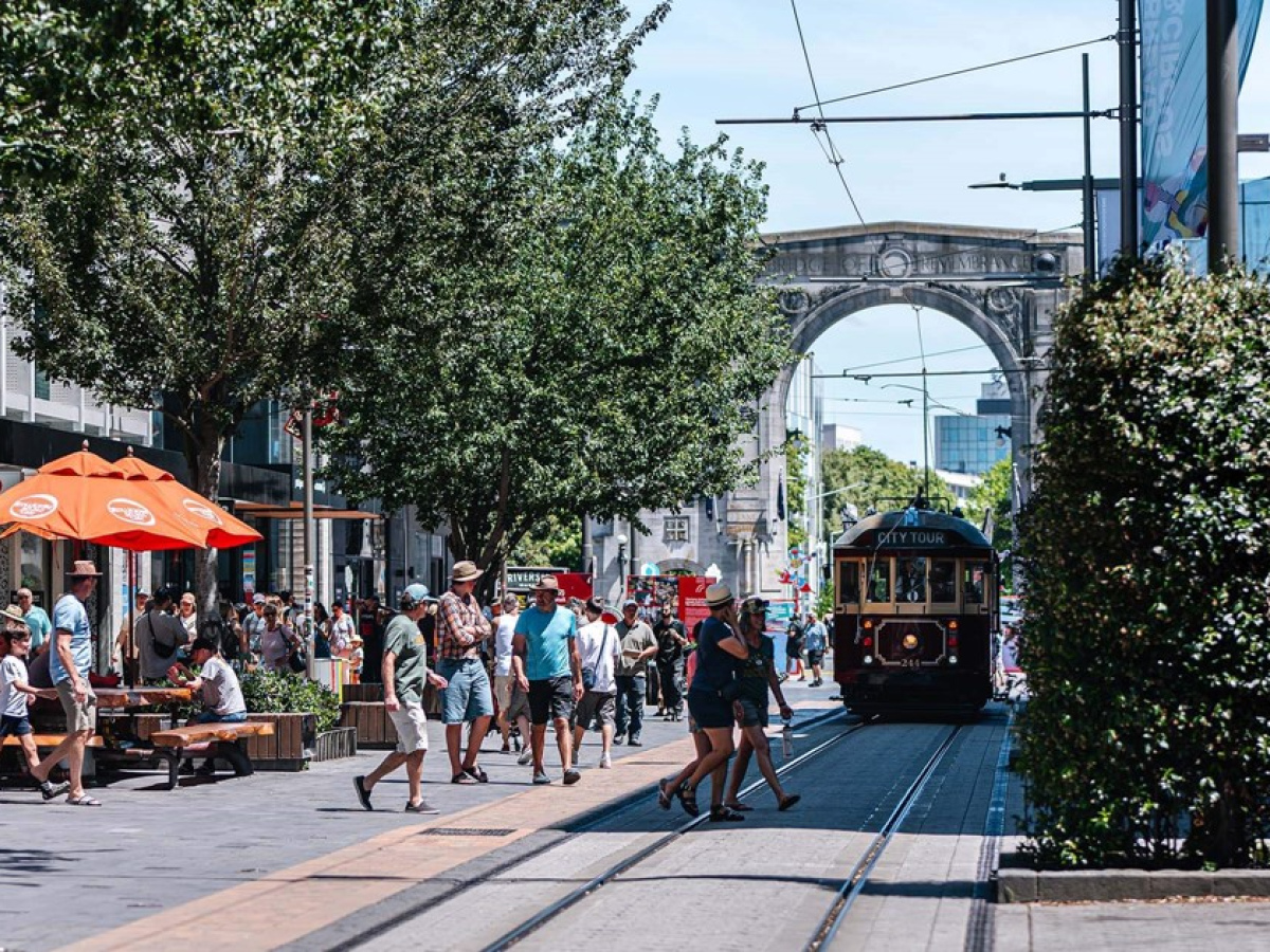 Cashel Street in the Christchurch city centre with the tram running through. The Bridge of remembrance is in the background and there are many people out shopping.