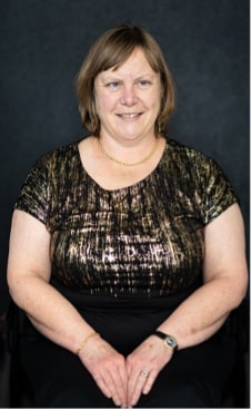 Genevieve McLachlan sitting in a relaxed pose, hands together in her lap, with smile on her face. She has chin length light brown hair and is wearing a fancy gold and black shirt.