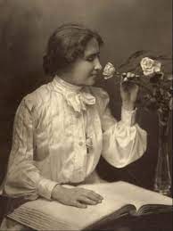 Helen Keller sitting with a large braille book open on her lap and she is smelling a rose that is being held in her left hand