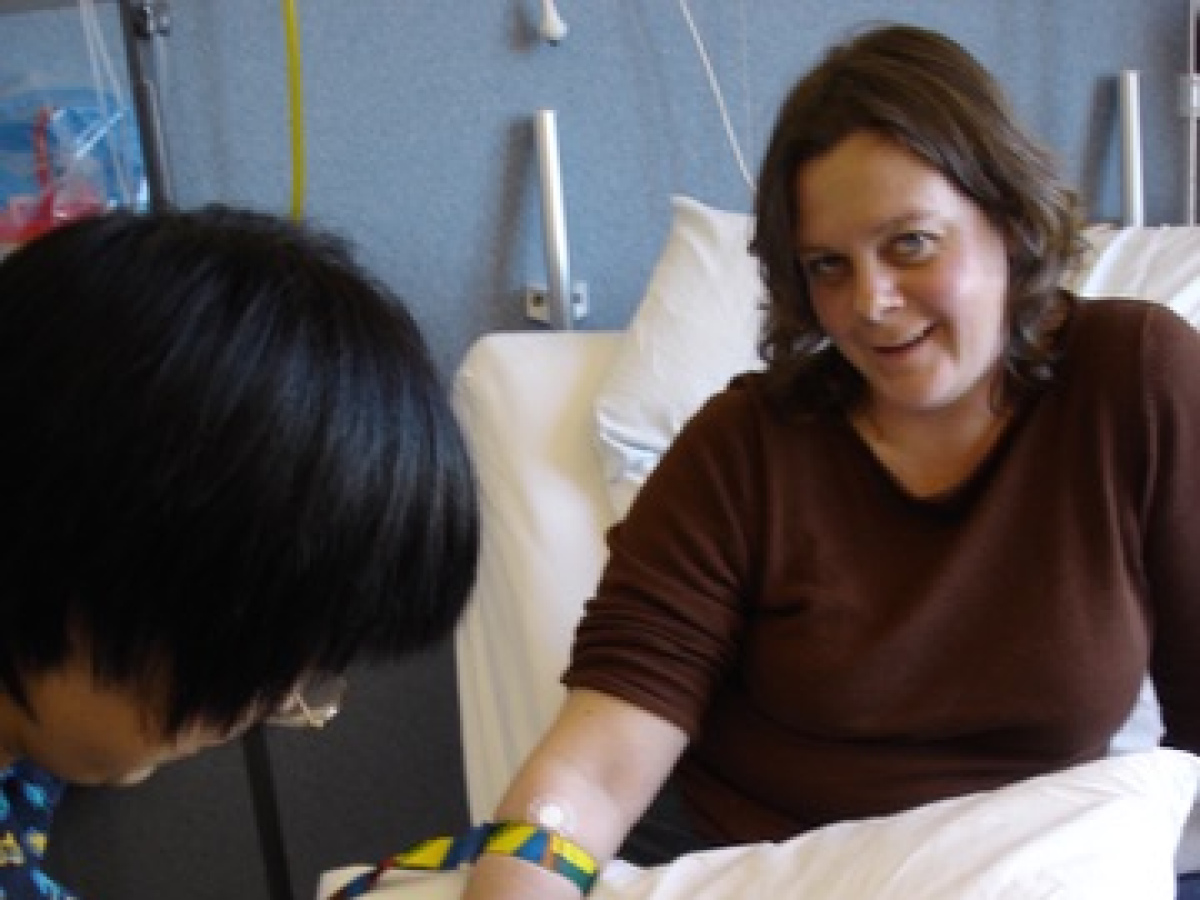 Chandra in hospital bed, her bandaged arm rests on a pillow, as a nurse is checking the arm.