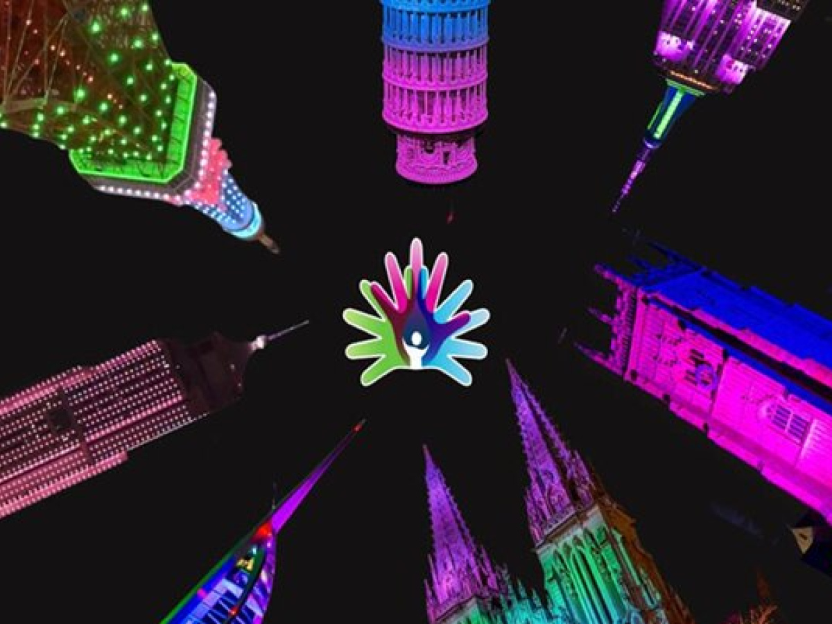 Colorfully illuminated landmarks arranged in a circular pattern against a black background, all pointing inward toward a multicolored hand-shaped Rare Disease Day logo at the center.