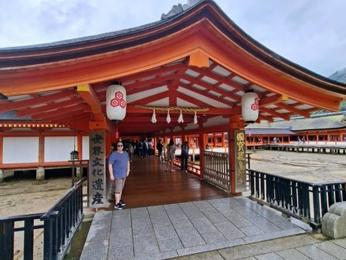 Katherine stands at the entrance of a traditional Japanese shrine on Miyajima Island. The building has bright orange wooden beams, a curved roof, and hanging lanterns. A covered walkway extends over shallow water, with several visitors walking further ins