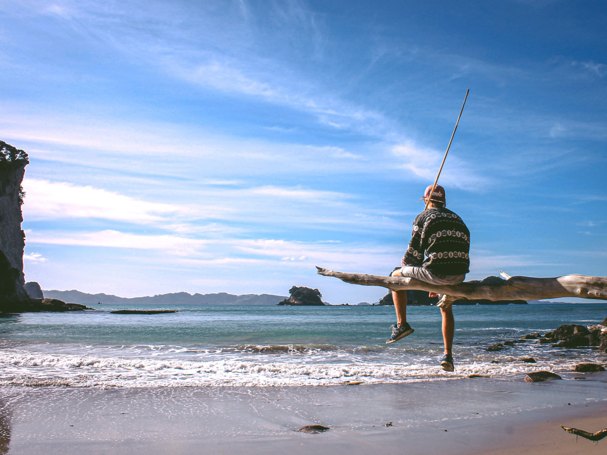 A young man sitting on a large piece of drift wood, looking out over the ocean.